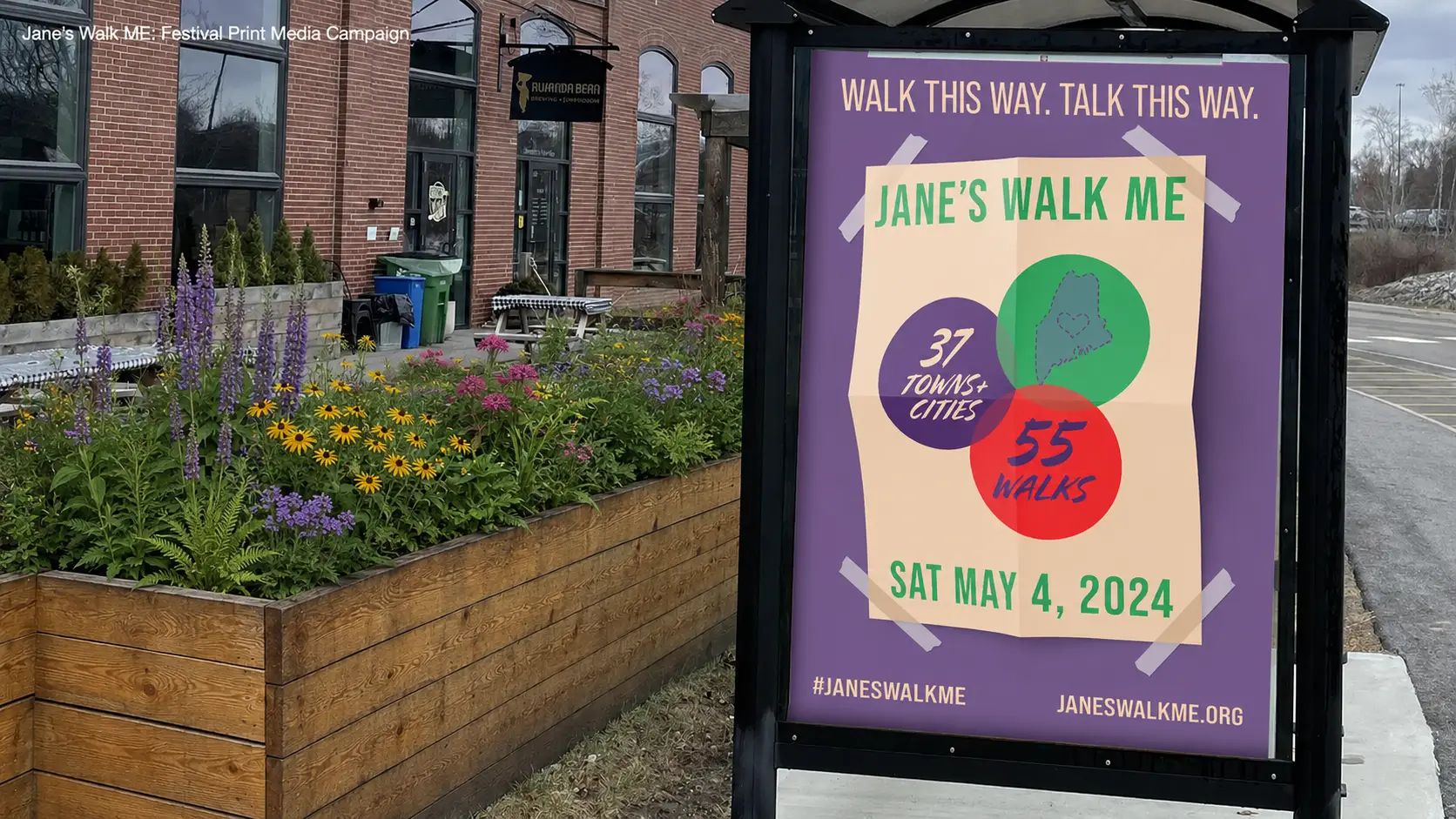 An outdoor photograph of a bus stop shelter, featuring a large purple poster on its back panel. The poster text reads - Janes Walk ME. The main section of the poster shows three interconnected circles: a green circle containing a small purple icon of the state of Maine with a heart, and a small purple circle on its left containing the text 37 TOWNS & CITIES. Intersecting the bottom of the green circle is a larger red circle with the text 55 WALKS. Below the circles, the date is stated as SAT MAY 4, 2024. The poster has light grey tape marks on all four corners. At the bottom right corner of the poster are the social media handle #JANESWALKME and the website JANESWALKME.ORG. In the foreground are large, weathered wooden planters containing dry plants.